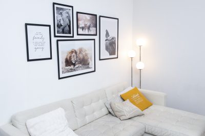 Kuvassa näkyy vaalea tekonahkainen sohva, jolla on sohvatyynyjä. Seinällä sohvan takana on taulukollaasi. Nukassa sohvan ja seinän välissä on kolmiosainen jalkalamppu.