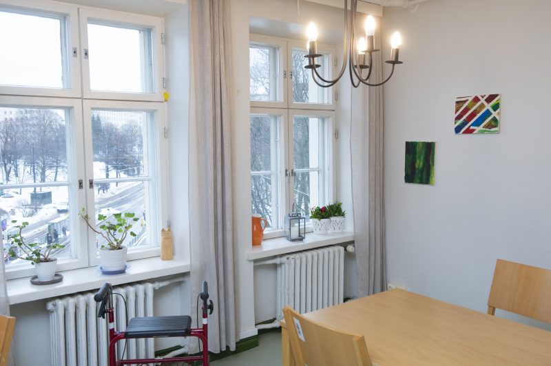Kuvassa näkyy huone, jonka ikkunoista näkyy talvinen kaupunkimaisema. Kuvassa näkyy myös puinen pöytä ja tuolit, rollaattori ja kaksi värikästä taulua seinällä.