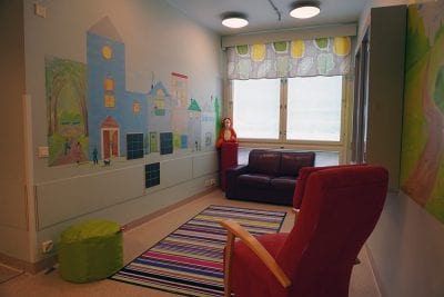 Kuvassa on toimipisteen olohuone, jossa on ikkunan edessä sohva, raidallinen matto lattialla ja punainen nojatuoli. Seinällä on lasten tekemä kuva, jossa on värikkäitä taloja.