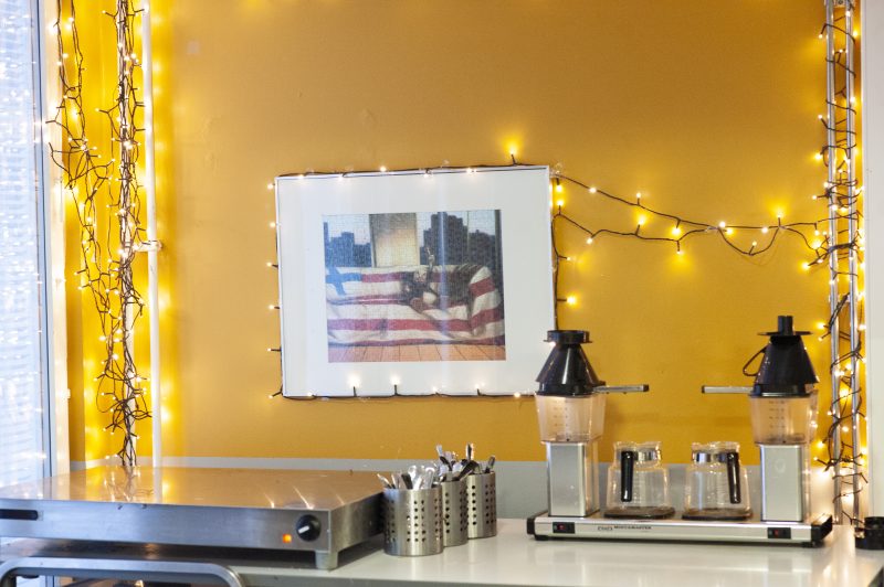 Lähikuva yhdestä Auroratalon kahvipisteestä. Pöydällä on kaksipannuinen kahvinkeitin, lusikoita ja kylmälevy maitoja varten. Pöydän takana oleva seinä on pirteän keltainen. Seinällä on taulu sekä jouluvaloja.