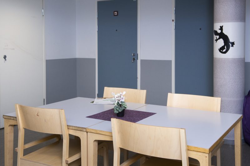 Sisäkuva Auroratalosta. Kuvassa näkyy neljän hengen pöytä, jolla pieni violetti pöytäliinaa ja pieni kukkiva viherkasvi. Takaseinällä on kaksi sinistä ovea.