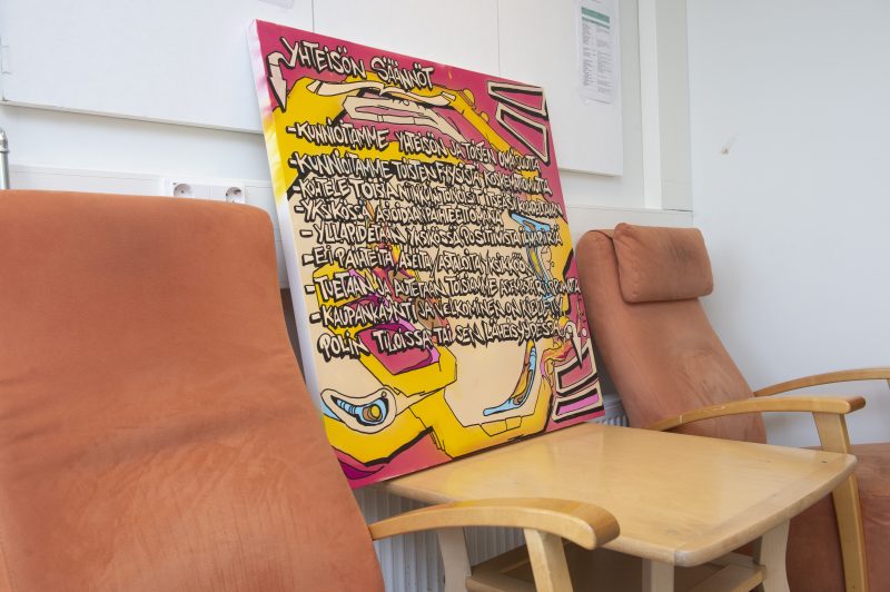 Sisäkuva Breikki Helsingistä. Lähikuvassa näkyy värikäs taulu, johon on kirjattu yhteisön säännöt. Taulu on sijoitettu pienelle pöydälle ja se nojaa seinää vasten kahden nojatuolin välissä.