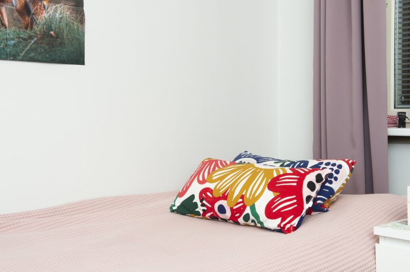 Sängyllä päiväpeiton päällä kaksi pientä sisustustyynyä. Seinällä näkyy pieni osa vihreäsävyisestä taulusta sekä roosan värinen verho.