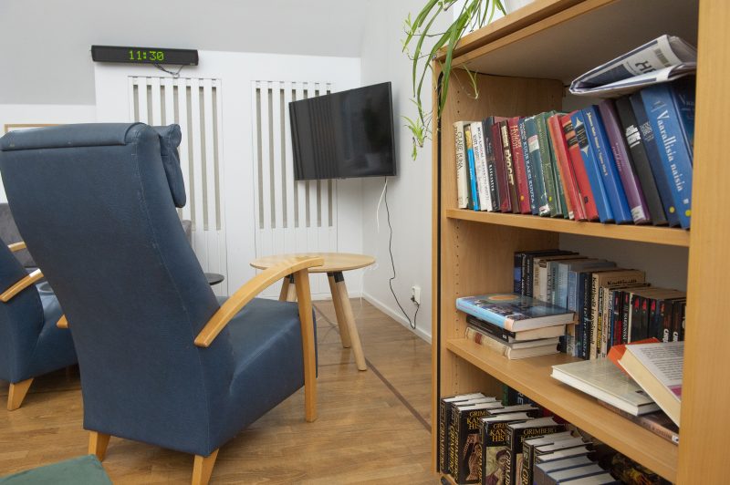 Yksityiskohtainen kuva toimipisteestä. Kuvan edustalla avonainen kirjahylly, jossa on kirjoja. Huoneen nurkassa on seinään kiinnitetty televisio. Oleskelutilassa näkyy myös kaksi sinistä nojatuolia sekä pieni sohvapöytä.