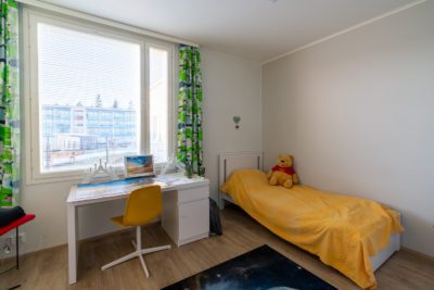 Kuvassa on lapsen huone. Sängyssä on keltainen päiväpeitto ja Nalle Puh -pehmolelu. Ikkunassa on kuviolliset vihreät verhot. Ikkunan edessä on työpöytä ja sen edessä keltainen tuoli.