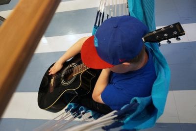 Kuvassa on lippispäinen lapsi kitara sylissään riippumatossa istumassa.