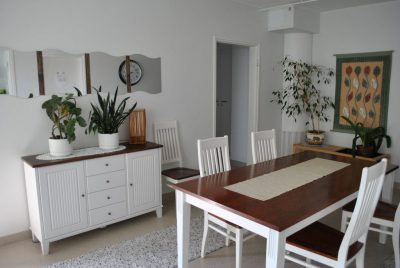 Sisäkuva Jokipolusta. Kuvassa näkyy neljänhengen ruokailuryhmä ja seinän vieressä oleva senkki. Senkin päällä on viherkasveja. Huonekalujen tasot ovat tummanpunertavaa puuta, ja muu viimeistely on valkoista. Seinällä on koristeena aaltopeili.