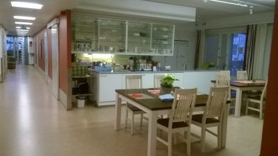 Sisäkuva Joukontalon ruokasalista. Kuvassa näkyy kaksi neljän hengen ruokapöytää, joiden takana on saareke ja keittiö. Sivussa näkyy myös käytävä muihin tiloihin.