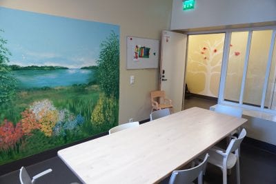 Sisäkuva yksikössä- Kuvassa on huone, jossa on ruokapöytä ja sen ympärillä tuoleja. Seinällä on suuri maisemakuva. Avoimesta ovesta näkyy käytävä, jonka seinällä on maalaus jossa on puu ja lintuja.