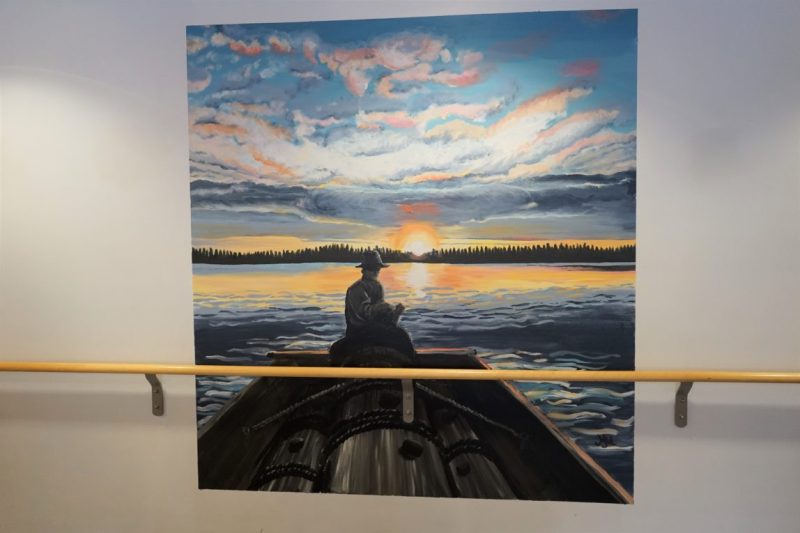 Seinällä taulu. Taulussa on maalaus henkilöstä veneessä auringon noustessa.