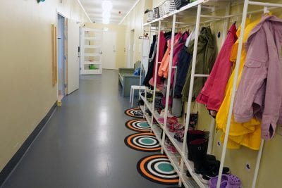 Sisäkuva toimipisteestä. Kuvassa käytävä, jossa keltainen seinä ja harmaa lattia, oikealla puolella kuvaa vaatenaulakko jossa on värikkäitä vaatteita ja pyöreitä raidallisia mattoja.