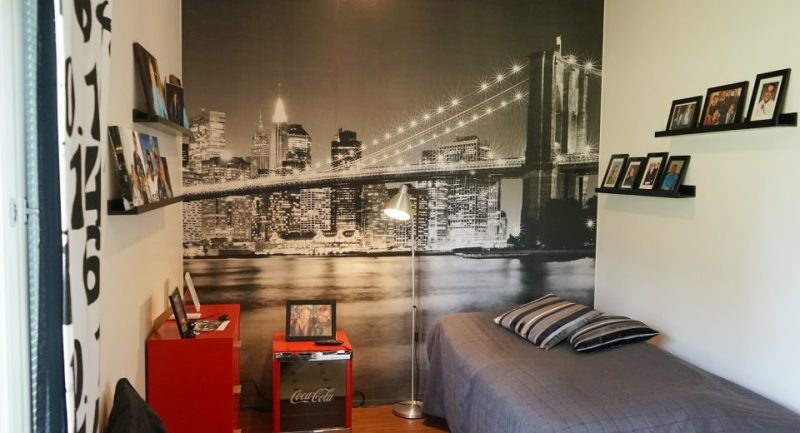 Kuva Kompassin asuinhuoneesta. Huoneessa on sänky, jolla on harmaa päiväpeitto sekä punaiset yöpöytä ja lipasto. Seinällä on tauluhyllyjä, joilla on kuvia. Takaseinällä on San Fransiscon kaupunkimaisema -tapetti.