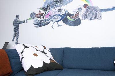 Kuvassa näkyy sininen sohva, jolla on unikkokuvioinen koristetyyny. Seinällä sohvan takana on maalaus, jossa nuorimies suihkuttaa spray-maalipullosta mm. riikinkukon, kalan, kolibrin, sateenkaaren ja koiran.