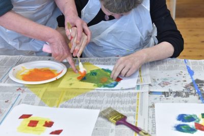 Kuvassa seisova henkilö auttaa istuvaa henkilöä maalaamaan värikkäillä maaleilla. Pöydällä on sanomalehtipaperia suojana.