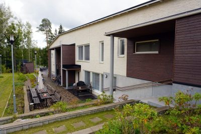 Ulkokuva Lastutieltä. Rakennus on vaalean sävyinen ja siinä on tummapuisia yksityiskohtia. Pihalla on puutarhakalusteina neljä pöytäryhmää, joista kolme ovat vierekkäin ja kahden aurinkovarjon suojaamia. Talon vierustalla on polkupyöriä. Pihapiiri on vehreä.