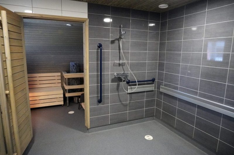 Lastutien suihku- ja saunatilat. Kuvassa näkyy tyylikäs harmaalaattainen suihkutila, jossa on mustia tukitankoja. Ovi on auki saunaan.