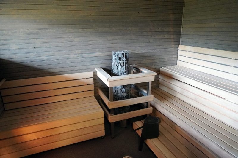 Lastutien sauna. Kiuas on turvallisesti aidattu ja sen molemminpuolin on lauteet. Toisen puolen lauteet ovat kolmitasoiset ja toinen puoli on penkkimallinen