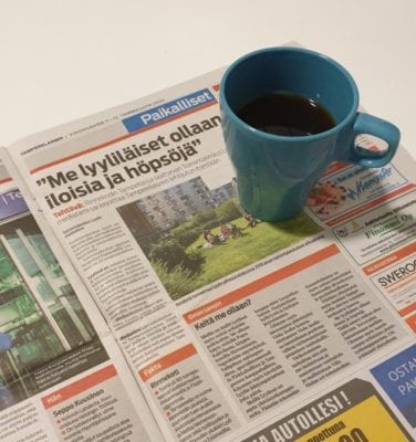 Kuvassa on sanomalehti jonka päällä on turkoosi kahvikuppi. Sanomalehdessä on juttu toimintakeskus Lyylistä, jonka otsikossa lukee "Me lyyliläiset ollaan iloisia ja höpsöjä".