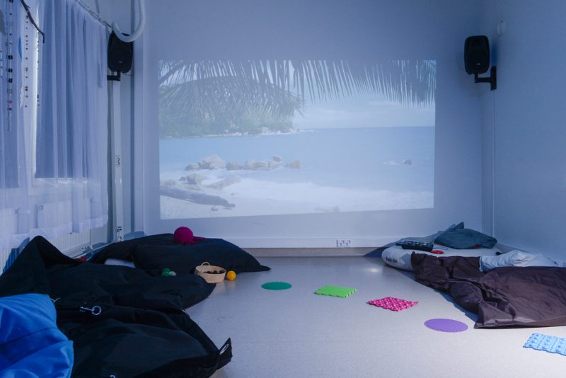 Kuva huoneesta, jonka takaseinälle heijastettu kuva rantamaisemasta. Lattialla tummia säkkituoleja sekä pieniä värikkäitä askellusalustoja.