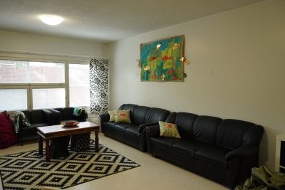 Kuvassa on toimintakeskus Meripihkan olohuone, jossa on kaksi mustaa sohvaa ja kaksi nojatuolia, mustavalkoinen matto ja sen päällä puinen pöytä. Seinällä on värikäs taulu. Nojatuolien takana on ikkuna.