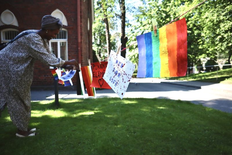 Nainen ripustaa taideteoksia pyykkinarulle ulkona. narulla roikkuu myös sateenkaarilippu.