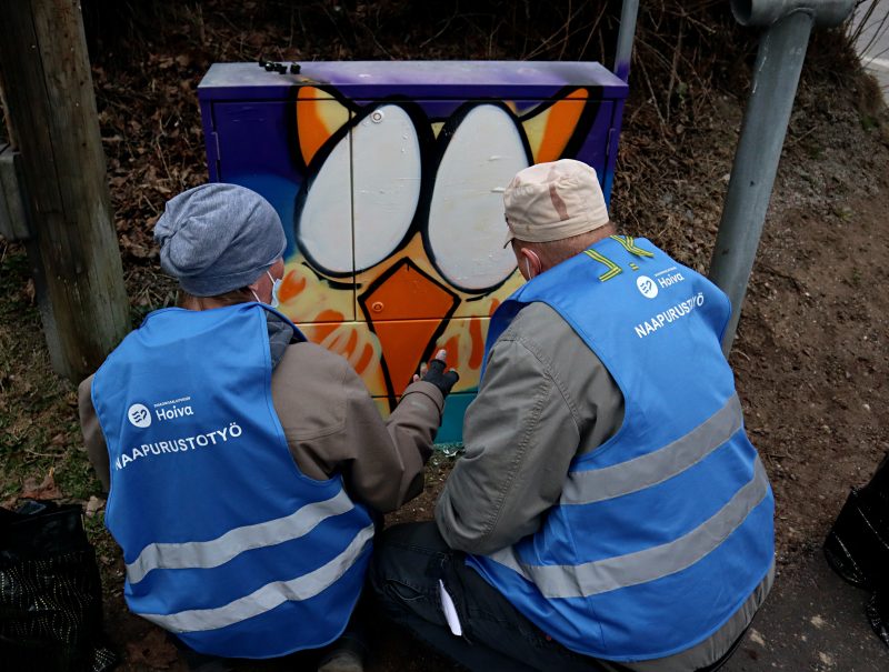 Kaksi ihmistä sininen huomioliivi päällä maalaa sähkökaappia värikkääksi. 