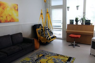 Nyyttipuiston olohuoneessa näkyy sohva, nojatuoli sekä piano. Seinällä on keltainen taulu ja matossa on keltaharmaa kuviointi.