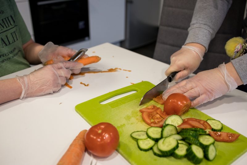 Kahden henkilön kädet näkyvät kuvassa, molemmilla on kertakäyttöiset kumihanskat käsissään. Toinen kuorii porkkanaa, toinen pilkkoo tomaatteja ja kurkkua vihreän leikkuulaudan päällä.