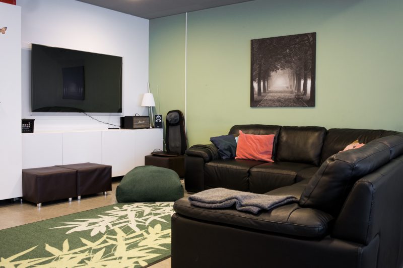 Kuvassa tv-nurkkaus, jossa on musta kulmasohva, jonka päällä on punainen tyyny. Huoneessa on vihreä seinä ja vihreä matto, television alla on valkoinen lipasto, jonka päällä on äänentoistolaite ja valaisin.