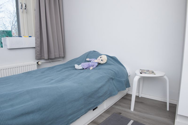 Paletin asuinhuonekuvassa näkyy sänky, jolla on sininen päiväpeitto ja nukke. Sängyn vieressä on yöpöytä, jolla on lehti. Ikkunalla on harmaat verhot.
