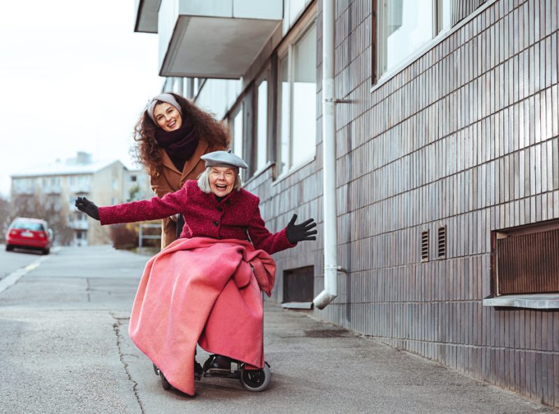 Hoitaja työntää pyörätuolissa istuvaa mummoa alamäkeen, hymyilevät kasvot