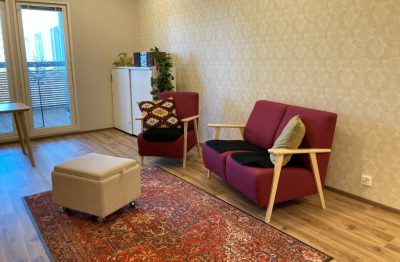 Säteen tv-huoneessa on viininpunainen sohva ja nojatuoli. Jalkojen ulottuvilta löytyy pyörillä kulkeva beige rahi ja lattialla on punainen matto. Takanurkasta löytyy lipasto, jonka päällä on radio ja köynnöskasveja.