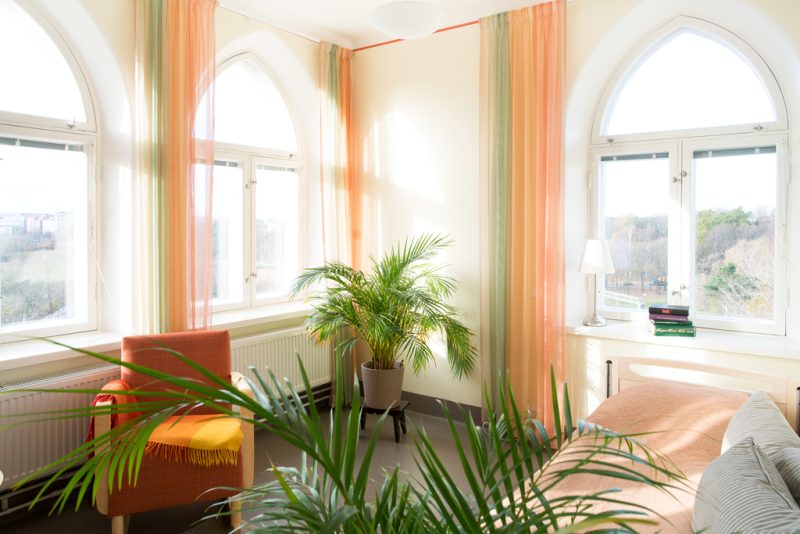Kuvassa on asukkaan huone, jossa on kolme vanhaa kaari-ikkunaa kahteen ilmansuuntaan. Niistä tulee valoa sisään. Vaaleassa huoneessa on oranssisävyiset verhot ja viherkasveja. Oranssilla nojatuolilla on viltti, sängyllä päiväpeitto ja kaksi tyynyä.