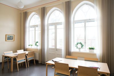 Kuvassa on toimipisteen ruokasali, jonka takaseinällä on kolme vanhaa kaari-ikkunaa. Leveillä ikkunalaidoilla on viherkasveja, verhot ovat vaaleat. Kahta puista ruokapöytää kiertää tuolit.