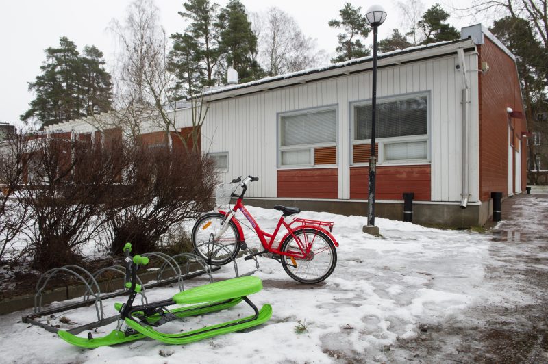 Talvinen kuva rakennuksesta ulkoa. Pyörätelineessä kiinni yksi punainen polkupyörä sekä neonvihreä rattikelkka.