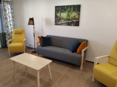 Tuulikanteleen olohuoneessa on harmaa sohva ja kaksi keltaista nojatuolia sekä sohvapöytä. Seinällä on metsäaiheinen taulu.