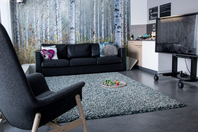 Toimipisteen tv-nurkkaus. Kuvassa on musta sohva ja nojatuoli, lattialla on harmaasävyinen pehmeä matto. Sohvan takana seinällä on koivikko-tapetti.