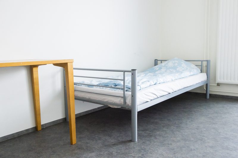 Kuva Wäylän asuinhuoneesta. Kuvassa näkyy pedattu sänky ja puinen pöytä. Seinät ovat valkoiset ja lattia harmaa.