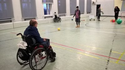Toimintakeskus Kartanonhakan liikuntasalo, jossa neljä ihmistä ja palloja. Yksi ihmisistä istuu pyörätuolissa.