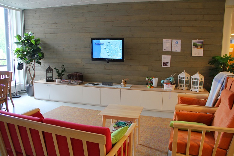 Oleskelutilat toimipisteessä. Oleskelutilassa on oranssin ja pinkin värisiä tuoleja ja sohvia sekä puun sävyinen pieni pöytä. Yhdellä seinustalla on seinään kiinnitetty televisio sekä koko seinän mittainen matala kaapisto. Kaapiston päällä on erilaisia koriste-esineitä ja viherkasveja.