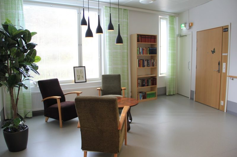 Vaaleasävyinen kuva oleskelutilasta, jossa kolme nojatuolia ja pöytä, korkea viherkasvi, kirjahylly sekä ovi toiseen tilaan. Seinällä isot ikkunat, joiden sivuilla vaaleanvihreät ohuet verhot.