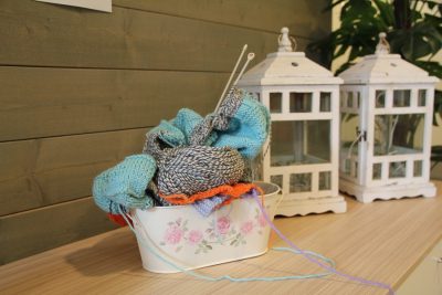 Kuvassa on käsityökori, jossa on kukkakuvioita ja jossa on päällä lankakeriä ja neulepuikkoja. Korin vieressä on kaksi valkoista lyhtyä.