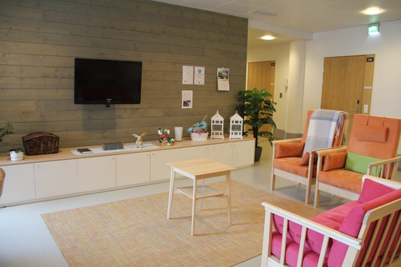 Oleskelutilat toimipisteessä. Oleskelutilassa on oranssin ja pinkin värisiä tuoleja ja sohvia sekä puun sävyinen pieni pöytä. Yhdellä seinustalla on seinään kiinnitetty televisio sekä koko seinän mittainen matala kaapisto. Kaapiston päällä on erilaisia koriste-esineitä ja viherkasveja.