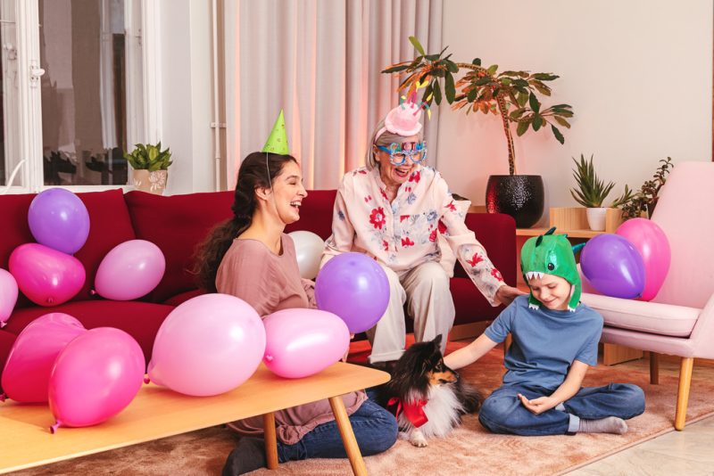 Huoneessa kolme ihmistä värikkäissä paperihatuissa, ympärillä ilmapalloja, lattialla mustavalkoinen koira.