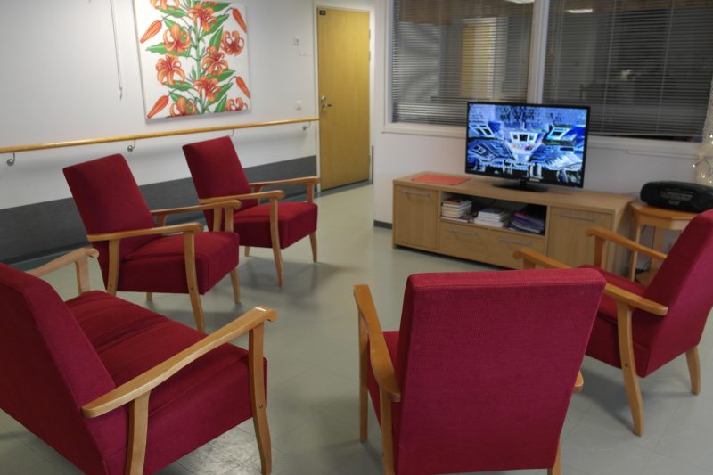 Sisäkuva toimipisteestä. Oleskelutilan keskellä punaisia nojatuoleja, tv-taso ja televisio.