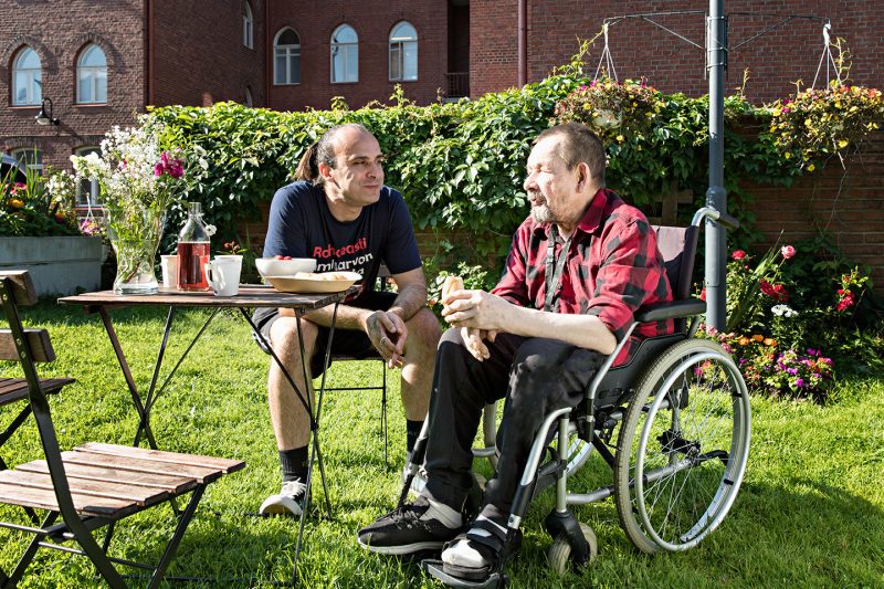 Hoitaja ja pyörätuolissa istuva asiakas nauttivat eväitä kesäisessä puutarhassa.