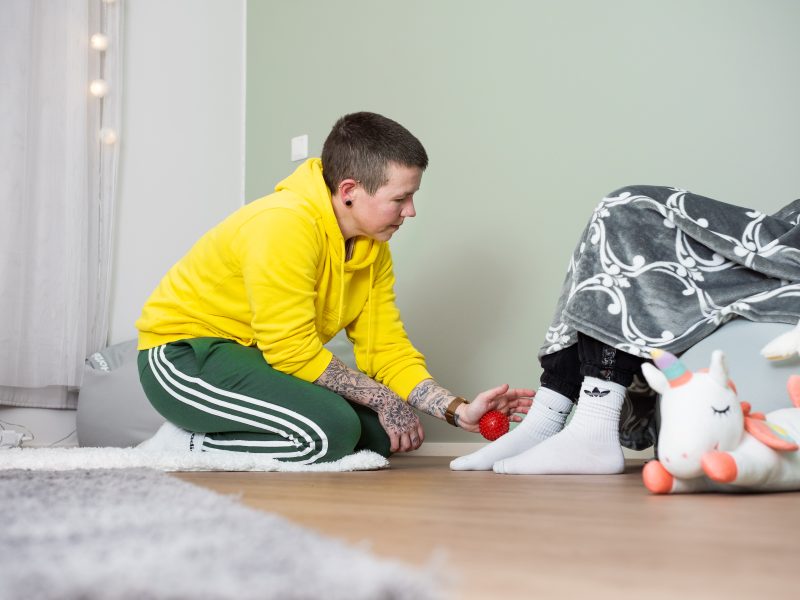 Keltaiseen huppariin pukeutunut nainen on polvillaan maassa ja hieroo tuolissa istuvan henkilön jalkaa nystyräpallolla. Tuolissa istuvalta näkyvät vain jalat.