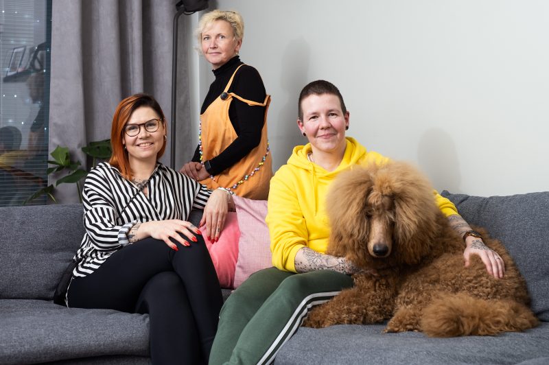 Kolme naista istuu sohvalla suuren ruskean koiran kanssa ja hymyilevät kameralle.