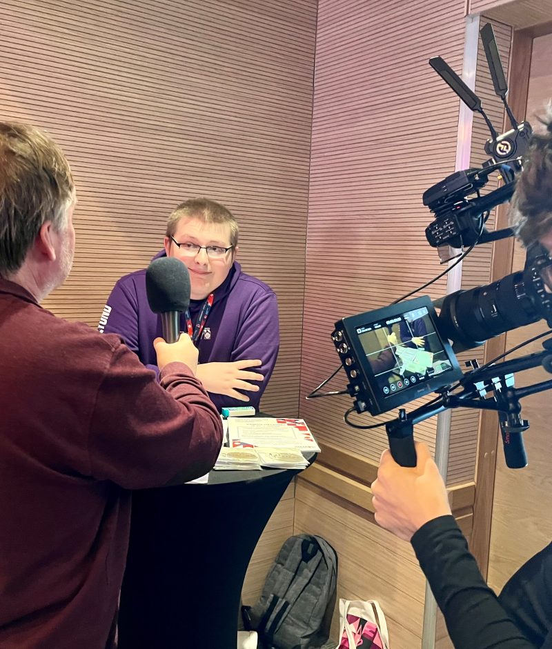 Nuori mies nojaa pystypöytään, toinen pitelee selkä kameraan päin mikrofonia, vieressä näkyy kamera, joka kuvaa haastattelua. 