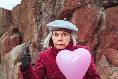 Baskeripäivän ikääntynyt nainen näyttää kameralle peukkua ja pitää kädessään vaaleanpunaista ilmapalloa.